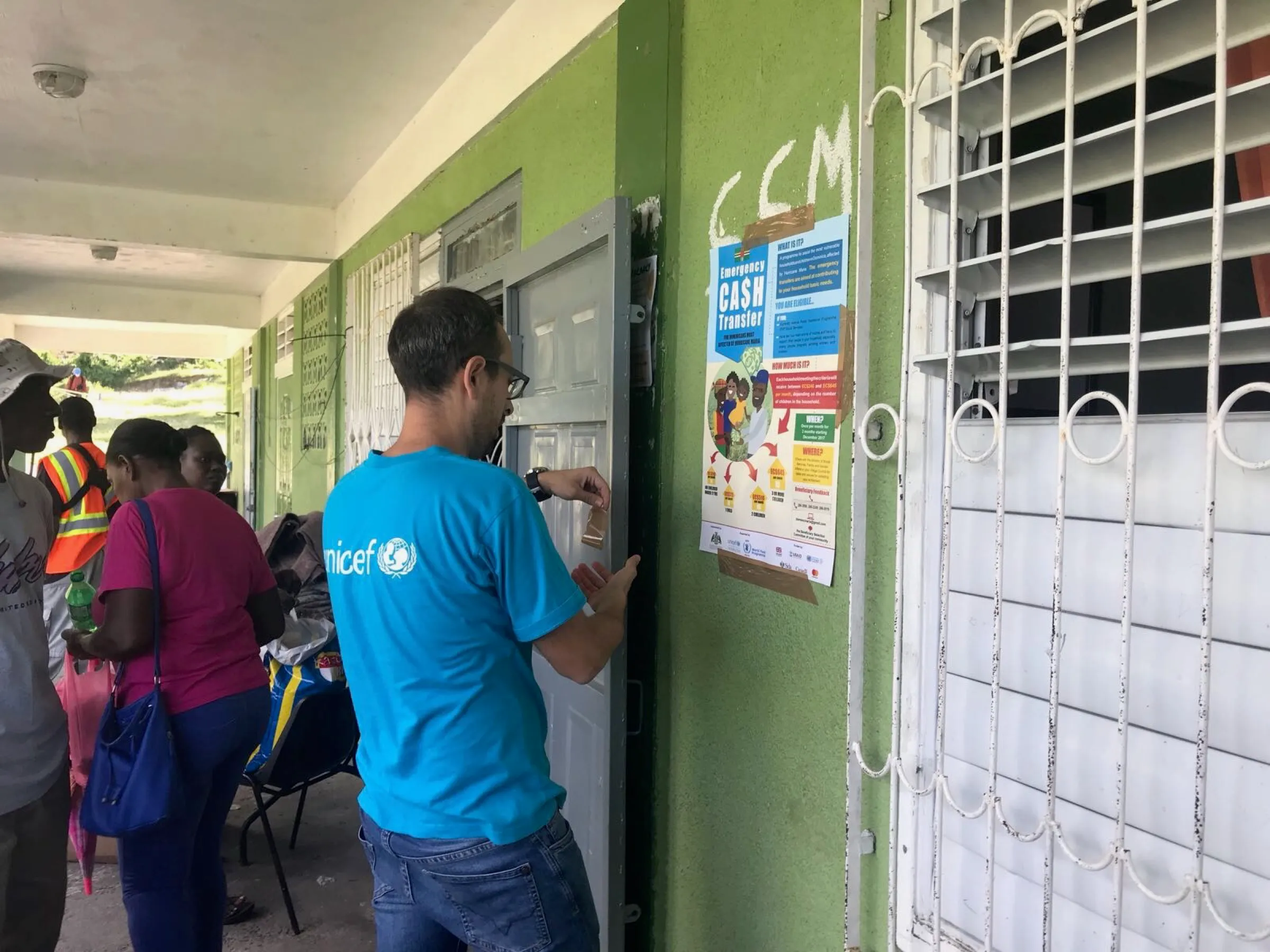 An Emergency CASH Transfer information poster being affixed to a community building entrance in the Eastern Caribbean, with people arriving.