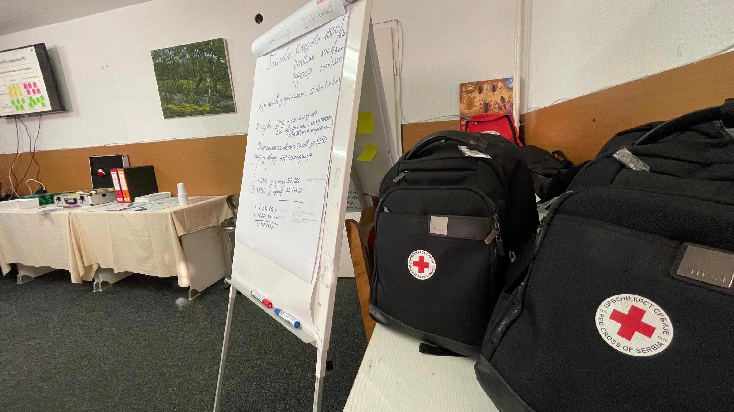 A Red Cross of Serbia training session: a flip chart with handwritten Cyrillic calculations next to two Red Cross of Serbia branded equipment bags, with workshop tables and documents in the background.