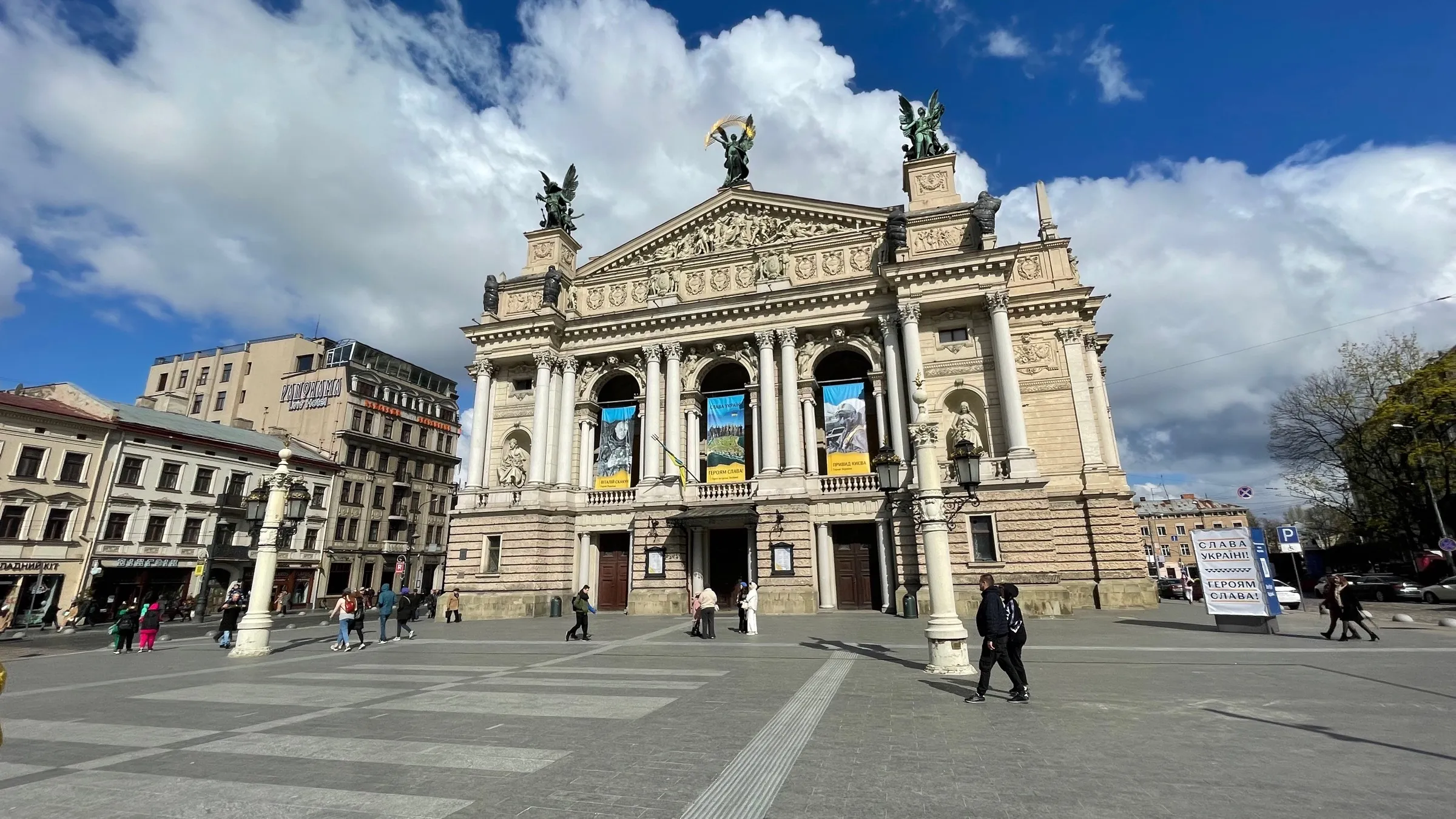 Lviv Opera House on a clear day in April 2022, with 'Slava Ukraini' banners flanking the entrance and civilians crossing the plaza in front.
